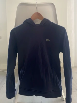 Mörkblå hoodie från Lacoste Sport - Snygg mörkblå hoodie från Lacoste Sport med klassisk krokodillogga på bröstet. Hoodien har huva och känguruficka framtill. Tillverkad i mjukt material som passar perfekt för en avslappnad stil.