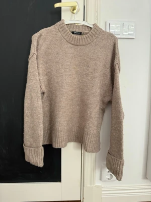 Beige stickad tröja med ribbade muddar - Mysig beige stickad tröja med rund halsringning och breda ribbade muddar vid ärmslut och nederkant. Tröjan har en avslappnad passform och är perfekt för kyliga dagar. Materialet ser ut att vara en mjuk ull- eller ullmix.