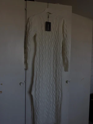 Vit lång stickad klänning - Lång, vit stickad klänning med kabelstickat mönster och lång ärm. Klänningen har en hög hals och är gjord i ett mjukt material som passar perfekt för kyligare dagar. Enkel och stilren design som ger en snygg siluett.