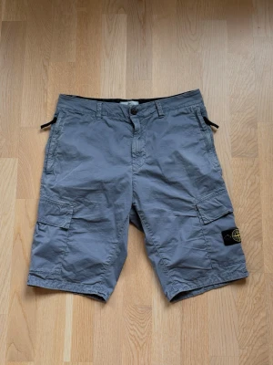 Grå cargoshorts med fickor från stone Island  - Snygga grå cargoshorts med flera praktiska fickor och knappstängning. Shortsen har en avslappnad passform och bälteshällor i midjan. Perfekta för varma dagar och har en klassisk look med patchdetalj på benet. Passar perfekt för både kvinnor och män! Originellt pris är 3999kr men säljer för 820!