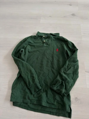 Grön långärmad henley med krage - Mörkgrön långärmad henley med krage och två knappar framtill. Skjortan har en röd broderad logga på bröstet och är tillverkad i ett bomullsliknande material. Perfekt för en avslappnad och stilren look.