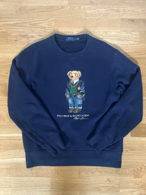 Blå sweatshirt Polo Bear Ralph Lauren - Mörkblå sweatshirt från Ralph Lauren med den ikoniska Polo Bear-trycket på bröstet. Rund halsringning, långa ärmar och ribbade muddar. Tillverkad i mjukt bomullsmaterial för en skön och avslappnad look. Perfekt för dig som gillar klassisk streetwear med en twist. Skriv gärna om ni har frågor mm, priset kan diskuteras vid rimliga bud.