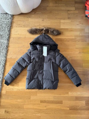 Grå Canada goose dunjacka med pälskantad huva  - Snygg grå Canada goose dunjacka med stor huva och avtagbar pälskant. Jackan har svarta muddar vid ärmsluten, två framfickor med dragkedja och en patch på ena ärmen. Perfekt för kalla vinterdagar och riktigt varm tack vare dunfyllningen.