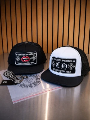 Chrome Hearts truckerkepsar svart/vit - Två Chrome Hearts truckerkepsar, en helsvart med broderade röda läppar och en svartvit med klassisk CH-logga. Båda har mesh på sidorna och justerbar snapback. Tuffa detaljer och tydliga broderier framtill. Perfekt för dig som gillar streetwear och statement-plagg.