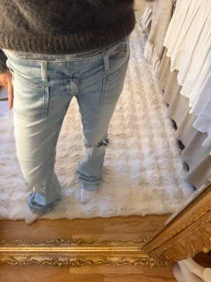 Ljusblå slitna bootcut jeans - Säljer ett par ljusblå jeans med bootcut passform och slitningar på både fram- och baksida. Jeansen har stora fickor fram och bak samt detaljerade sömmar. Perfekta för en avslappnad och trendig look.