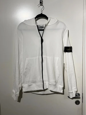 Vit hoodie från Stone Island - Snygg vit hoodie från Stone Island med dragkedja framtill och klassisk logotyp-patch på vänster ärm. Hoodien har huva, känguruficka och ribbade muddar. Perfekt för en clean och stilren look.