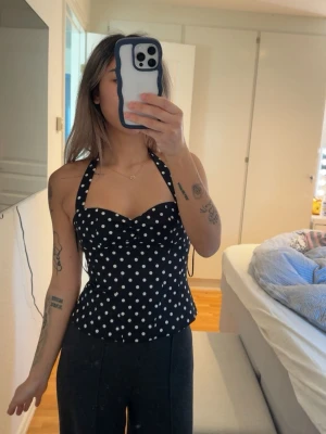 Svart prickig halterneck toppl - Snygg svart halterneck topp med vita prickar och söt sweetheart-urringning. Toppen har figurnära passform och knyts i nacken, vilket ger en retro och trendig vibe. Perfekt att styla med både jeans och kjol för en cool look.