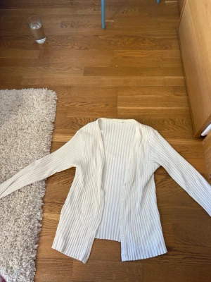 Vit ribbad kofta med knappar - Säljer en stilren vit kofta med ribbad struktur och knäppning framtill. Koftan har långa ärmar och en rak passform som passar till många olika outfits. Perfekt för lager på lager och enkel att matcha.