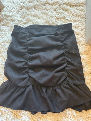 Svart volangkjol från Gina Tricot - Säljer en svart kortkjol från Gina Tricot med snygga volangdetaljer och rynk framtill. Kjolen har en elastisk midja och är gjord i ett lätt, strukturerat tyg som ger en cool look. Perfekt att styla med en crop top eller stickad tröja.