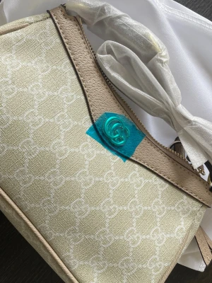 Beige handväska med GG-mönster - Snygg beige handväska från Gucci med klassiskt GG-mönster i vitt och detaljer i ljusbeige läder. Väskan har guldfärgad GG-logga framtill och dragkedja upptill. Perfekt accessoar för dig som gillar lyxig och tidlös design.
