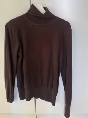 Brun polotröja i stickat material - Mysig brun stickad polotröja med lång ärm och ribbad krage. Tröjan har en klassisk passform och är tillverkad i ett mjukt, tunt material som känns skönt mot huden. Perfekt för lager på lager och enkel att matcha med olika outfits.
