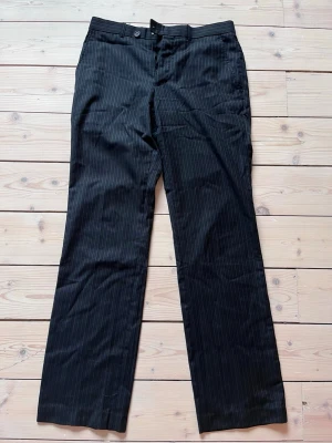 Svarta kostymbyxor pinstripe - Snygga svarta kostymbyxor med diskreta vita nålrandiga detaljer. Byxorna har raka ben och storlek M