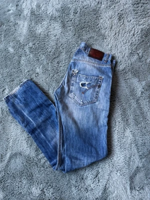 Blå slitna dondup jeans med hål och tvättade detaljer - Säljer ett par blå dondup jeans med slitningar, hål och tvättade detaljer. Jeansen har raka ben och klassisk femficksmodell. Materialet är denim i bomull och de har en cool, avslappnad look med slitna partier och färgskiftningar.