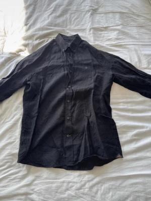 Ny Svart Morris Linneskjorta  - Säljer en ny svart skjorta i linne med klassisk krage och knappar framtill. Skjortan har lång ärm och en diskret broderad detalj på bröstet. Perfekt för en avslappnad men stilren look.