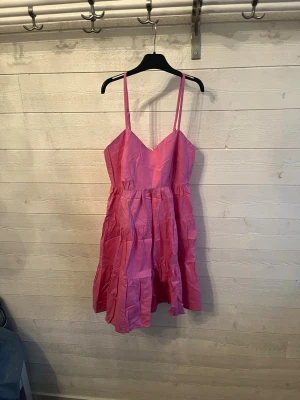 Rosa kort klänning från Sisters Point - Supersöt rosa klänning från Sisters Point med smala axelband och v-ringad urringning. Klänningen har en luftig, volangig kjol och är perfekt för varma dagar. Materialet känns lätt och somrigt, och modellen är kort och feminin.