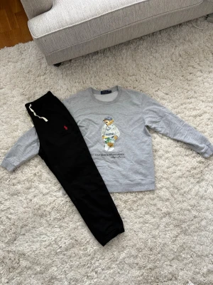 Loungewear set från Polo Ralph Lauren - Snyggt loungewear set med grå sweatshirt och svart byxa från Polo Ralph Lauren. Tröjan har en ikonisk Polo Bear-tryck framtill och rund halsringning. Byxan har dragsko i midjan och röd logga på benet. Perfekt för chill dagar hemma.