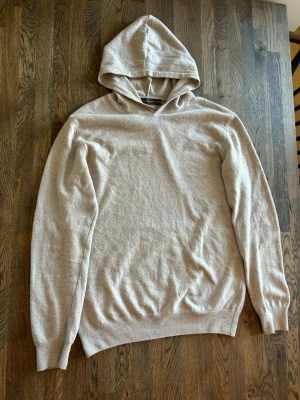 Beige merino hoodie från Oscar Jacobson - Snygg beige merino hoodie från Oscar Jacobson i mjukt och tunt material. Klassisk modell med huva och ribbade muddar vid ärmslut och nederkant. Perfekt för en clean och avslappnad stil.