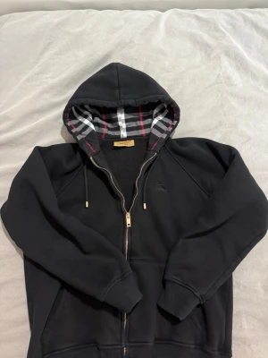Svart hoodie från Burberry med dragkedja M-L - Svart hoodie från Burberry med klassiskt rutigt foder i huvan i grått, vitt och rött. Dragkedja framtill, fickor på sidorna och guldfärgade detaljer på snörena. Tillverkad i mjukt material som känns skönt mot huden. Perfekt för en stilren och avslappnad look.