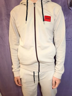 Beige hoodie med dragkedja från HUGO - Snygg beige hoodie från HUGO med dragkedja framtill, svart snörning i huvan och röd logotyp-patch på bröstet. Mjuk bomullskänsla och två fickor framtill. Perfekt för en chill och stilren look.