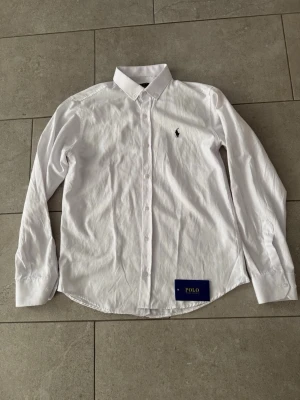 Vit skjorta från Polo Ralph Lauren - Klassisk vit skjorta från Polo Ralph Lauren med broderad logga på bröstet. Skjortan har lång ärm, knappar framtill och en stilren krage. Perfekt till både jeans och chinos för en clean look.