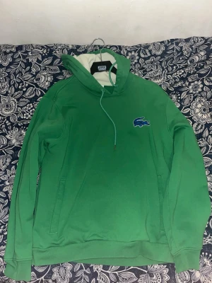 Grön hoodie från Lacoste med huva - Snygg grön hoodie från Lacoste med klassisk krokodillogga på bröstet. Hoodien har huva med dragsko och en mjuk, vit insida. Perfekt för en chill och sportig stil. Långa ärmar och fickor på sidorna.