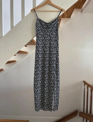 Blommig långklänning med smala band - Säljer en mörkblå långklänning med vita blommor och smala axelband. Klänningen har en snygg passform och v-ringad urringning framtill. Perfekt för varma dagar och enkel att styla med sneakers eller sandaler.
