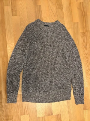 Gråmelerad stickad tröja - En klassisk gråmelerad stickad tröja med rund hals och långa ärmar. Tröjan har en grovstickad struktur och är perfekt för kyliga dagar. Passar dig som gillar en enkel och stilren look.