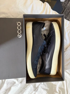 Mörkblå ECCO sneakers med vit sula - Snygga mörkblå sneakers från ECCO med klassisk låg profil och vit sula. Skorna har släta läderpaneler, matchande mörkblå snörning och diskret ECCO-logga på sidan. Perfekta för en stilren och avslappnad look.
