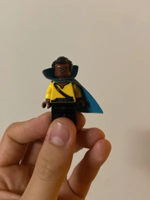 Lego minifigur med blå mantel - Säljer en Lego minifigur med gul tröja, svart byxor och en blå mantel. Figuren har lockigt svart hår och detaljerad tryck på överkroppen med bälte och symbol. Perfekt till samlingen eller som leksak.