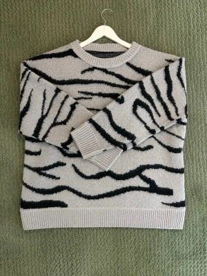 Grå stickad tröja med zebramönster - Snygg beige stickad tröja med svart zebramönster. Tröjan har rund halsringning, ribbade muddar vid ärmslut och nederkant samt en avslappnad passform.
