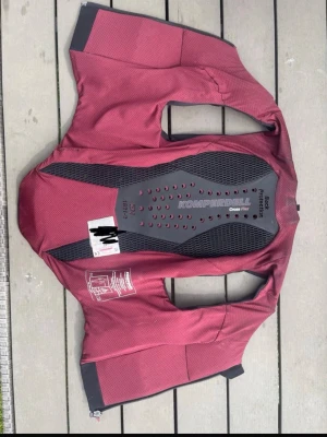 Komperdell ryggskyddsväst vinröd - Säljer en vinröd ryggskyddsväst från Komperdell, modell Cross Flex. Västen har svart skyddsplatta på ryggen med ventilationshål och dragkedja framtill. Perfekt för ridning, cykling eller andra sporter där extra skydd behövs.