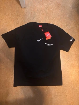 Svart Nike NOCTA x L'Art T-shirt - Svart t-shirt från Nike NOCTA i samarbete med L'Art de l'Automobile. Framsidan har vit Nike-logga och NOCTA-tryck, medan baksidan har ett stort gult bilmotiv och text. T-shirten har rund hals och tryck på båda ärmarna. Perfekt för dig som gillar streetwear.