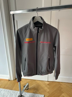 Grå vindjacka Prada Luna Rossa - Snygg grå vindjacka från Prada Luna Rossa med Pirelli-logga på bröstet. Jackan har hel dragkedja, två fickor med dragkedja och hög krage. Detaljer i rött och svart samt tryckta loggor framtill. Perfekt för dig som gillar sportig och stilren design. Modellen är 180 cm 