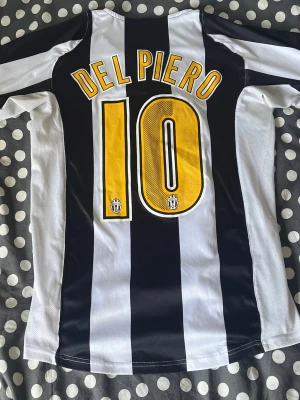 Juventus Del Piero matchtröja - Juventus kortärmad fotbollströja med svarta och vita ränder, Del Piero och nummer 10 på ryggen. Nike-logga, Juventus klubbmärke och två stjärnor på bröstet samt Sky Sport sponsortryck på framsidan. Perfekt för fans av klassisk fotboll.