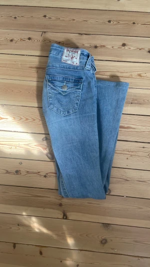 Blå bootcut jeans från True Religion - Säljer ett par klassiska blå bootcut jeans från True Religion. Jeansen har ikoniska sömmar och detaljerade bakfickor med lock och knapp. Tillverkade i mjukt denimtyg med en snygg tvätt och låg midja. Perfekta för en avslappnad och trendig look. De har ett litet hål på knät som är igen sytt med det syns inte