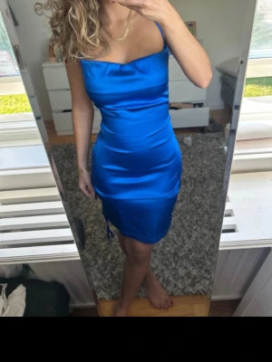 Blå satin klänning med snörning - Säljer en snygg blå kort klänning i satin med smala axelband och snörningsdetalj på sidan. Klänningen har en figurnära passform och en lätt glansig yta som ger en lyxig känsla. Perfekt för dig som vill sticka ut med en färgstark look.