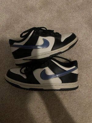 Svarta och vita Nike Dunk Low sneakers - Säljer ett par Nike Dunk Low sneakers i svart och vitt med blå Swoosh. Skorna har klassisk låg siluett, perforerad tåbox och svart snörning. Ovandelen är i läder och yttersulan är svart. Perfekta för dig som gillar streetwear och vill ha en clean look. Skorna har endast använts några gånger, och priset går att diskutera. 