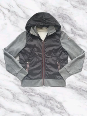 Moncler Cardigan Grå - Snygg Moncler Cardigan i grått och svart med huva och dragkedja framtill. Cardiganen har gråa ärmar i sweatshirtmaterial och svart glansig kropp. Två fickor med dragkedja och ribbade muddar. Perfekt för lager på lager under kyliga dagar. Storlek M men passar S. Köpt från Von Greven Closet