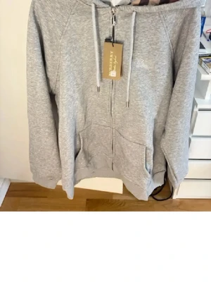 Ljusgrå zip hoodie med huva - En stilren ljusgrå hoodie med dragkedja framtill och två fickor. Hoodien har justerbar huva med snören och ribbade muddar vid ärmar och nederkant. Perfekt för en avslappnad och bekväm look. Materialet är mjukt och skönt, troligen bomullsblandning.