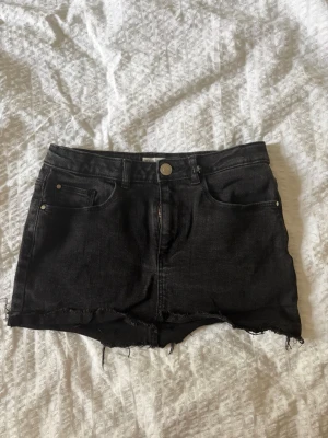 Svart jeanskjol med rå kant - Kort svart jeanskjol med fem fickor och rå, fransig kant nedtill. Kjolen har klassisk knapp- och dragkedjestängning samt bälteshällor i midjan. Perfekt för en avslappnad och trendig look. Storlek 158/164