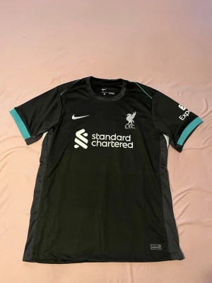 Liverpool FC svart matchtröja Nike - Snygg svart Liverpool FC matchtröja från Nike med vita och turkosa detaljer. Tröjan har klubbmärke och sponsortryck på bröstet samt logga på ärmen. Tillverkad i lätt och ventilerande funktionsmaterial, perfekt för träning eller match.