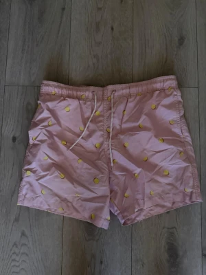 Rosa badbyxor med citronmönster - Säljer ett par ljusrosa badbyxor med broderade små citroner i gult. De har resårmidja med vit dragsko och en bakficka. Perfekta för stranden eller poolen och har en avslappnad passform. Materialet känns lätt och snabbtorkande.