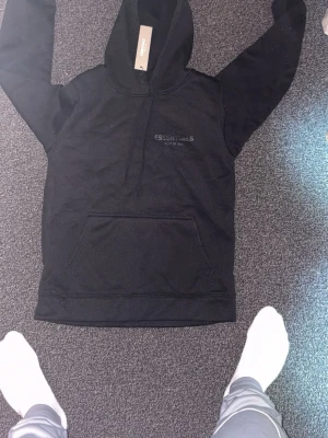 Svart Essentials Fear of God hoodie - Svart hoodie från Essentials Fear of God med stor logga på ryggen och mindre tryck på bröstet. Klassisk känguruficka framtill och huva. Perfekt för en avslappnad och trendig streetwear-look.