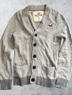 Hollister Cardigan Grå - Strl S - Säljer en stilren, snygg och sällsynt cardigan från Hollister i storlek S och grå färg. Plagget är i mycket bra skick (9/10) och redo för en ny ägare. Perfekt för dig som letar efter en stickad tröja med klassisk krage, mörkare grå detaljer och broderad logotyp från Hollister. Mått: Axel till axel / Bröst: 38 cm Längd: 66 cm Ärmlängd: 58 cm  Skickas snabbt och packas noggrant. Hör av dig vid frågor eller om du vill ha fler bilder!  