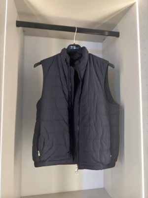 Mörkblå dunväst från Oscar Jacobson - Snygg mörkblå dunväst från Oscar Jacobson med hög krage och dragkedja framtill. Västen har två sidofickor med dragkedja och quiltad design. Perfekt att bära över en hoodie eller tröja för extra värme. 44=xs