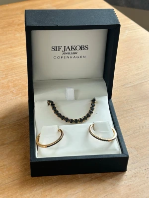 Oanvända Sif Jakobs örhängen + Caroline Svedblom armband - Aldrig använda. Har tyvärr inget kvitto då de var en present, men ni kan se gravyren på bilderna.  Armband: Caroline Svedblom (tror modellen Zara Bracelet), handgjort i guldpläterad mässing med Swarovski-kristaller. Nypris 800-900kr  Örhängen: SIF Jakobs (Ellera Grande Creolo). Tillverkade i 18k guldpläterat sterlingsilver med svarta cubic zirconia-stenar. Kommer i originalask. Nypris 1300-1400kr  Kan även sälja separat, kom med prisförslag