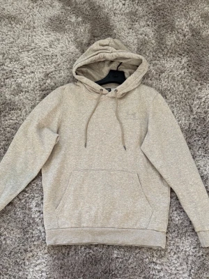 Beige hoodie från Under Armour - Snygg beige hoodie från Under Armour med broderad logga på bröstet. Klassisk känguruficka framtill, justerbar huva med dragsko och ribbade muddar vid ärmar och nederkant. Tillverkad i mjukt sweatshirtmaterial som känns skönt mot huden.