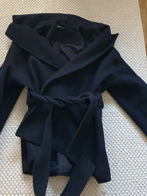 Svart kappa med knytskärp - Stilren svart kappa med bred krage och knytskärp i midjan. Kappan har en klassisk, rak passform och är tillverkad i ett mjukt tyg som ger en snygg siluett. Perfekt för dig som vill ha en enkel men trendig look.