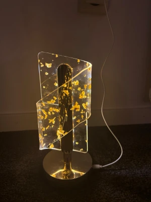 Modern LED bordslampa i akryl - Unik bordslampa med modern design i transparent akryl och guldfärgade detaljer. Lampan har en geometrisk form och står på en rund metallfot. Den lyser med ett varmt sken och ansluts via USB-kabel. Perfekt som dekorativ belysning i hemmet.