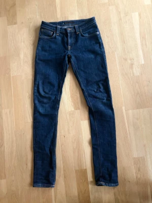 Mörkblå slim jeans från Nudie Jeans W27 L30 - Säljer ett par mörkblå slim jeans från Nudie Jeans med klassiska orange sömmar och läderpatch bak i midjan. Jeansen har fem fickor och är tillverkade i stretchigt denimtyg för en bekväm passform.